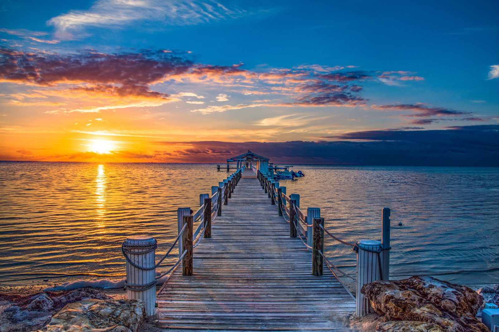 A wooden dock with rope railings leading out over calm water toward a bright orange sunset